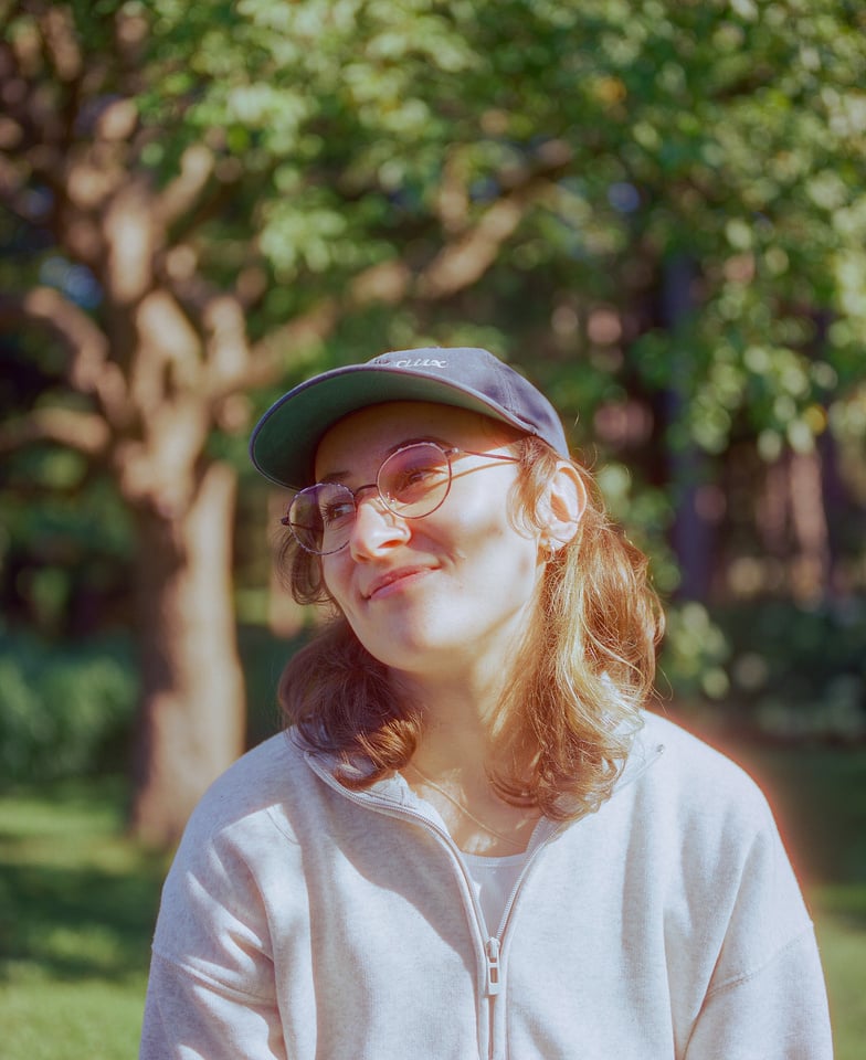A calm person smiling wearing glasses and a baseball cap in nature in the sun